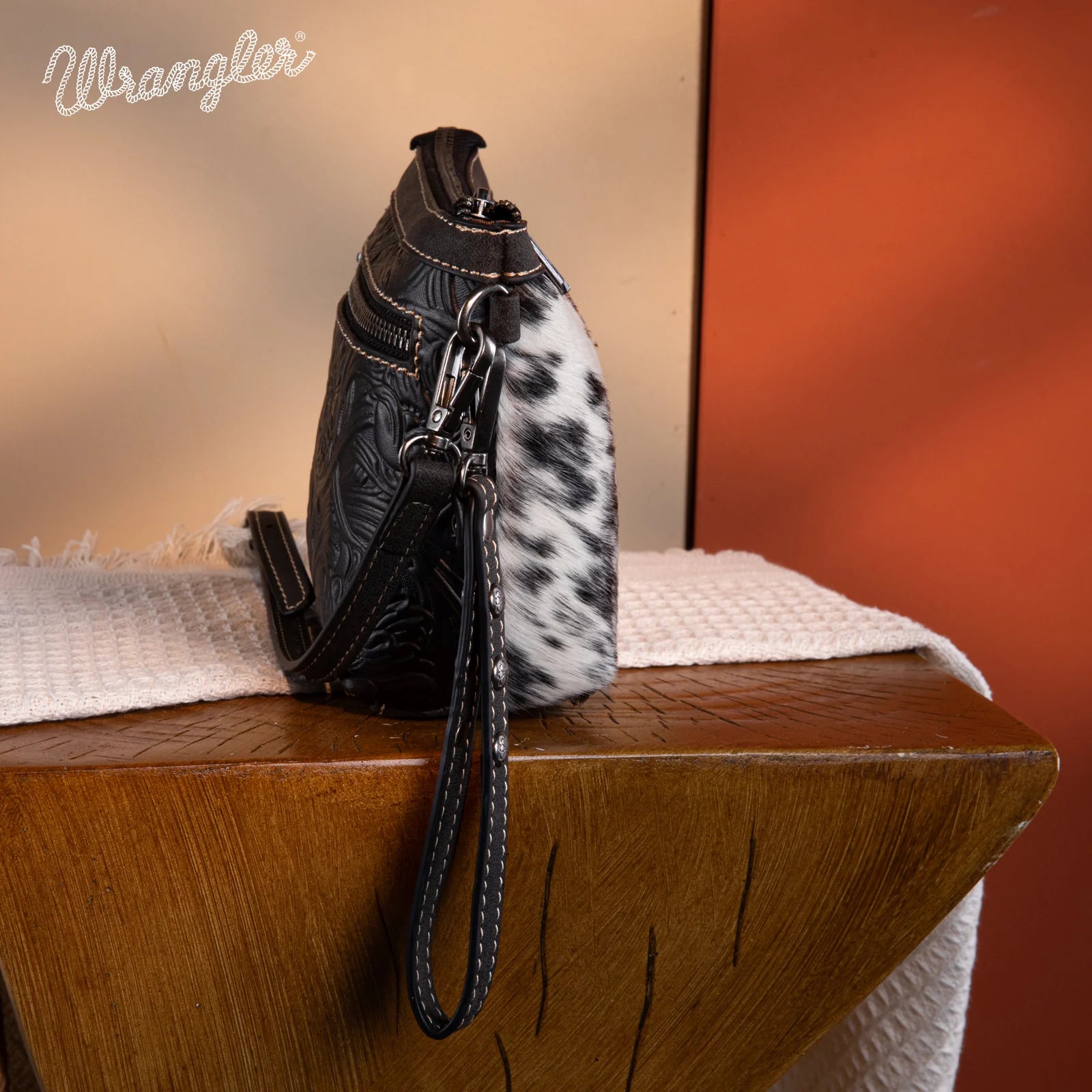 Side view of Wrangler Genuine Hair-On Cowhide Crossbody wristlet with genuine black and white cowhide, tooled black PU leather, and detachable studded wrist strap.