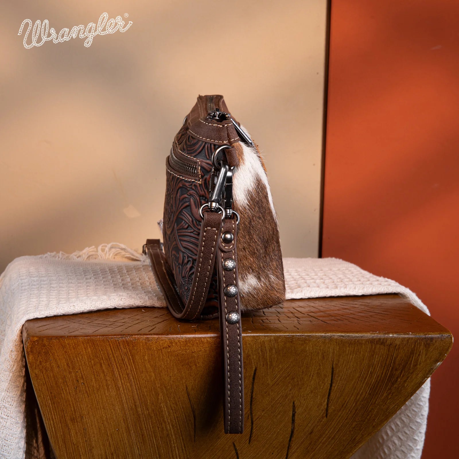 Side view of Wrangler wristlet with tooled PU leather, genuine hair-on cowhide panel, studded strap, and Western floral design.